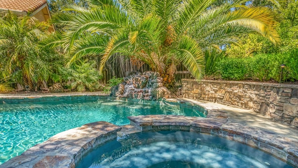 A swimming pool with a hot tub, surrounded by lush greenery and a stone wall.