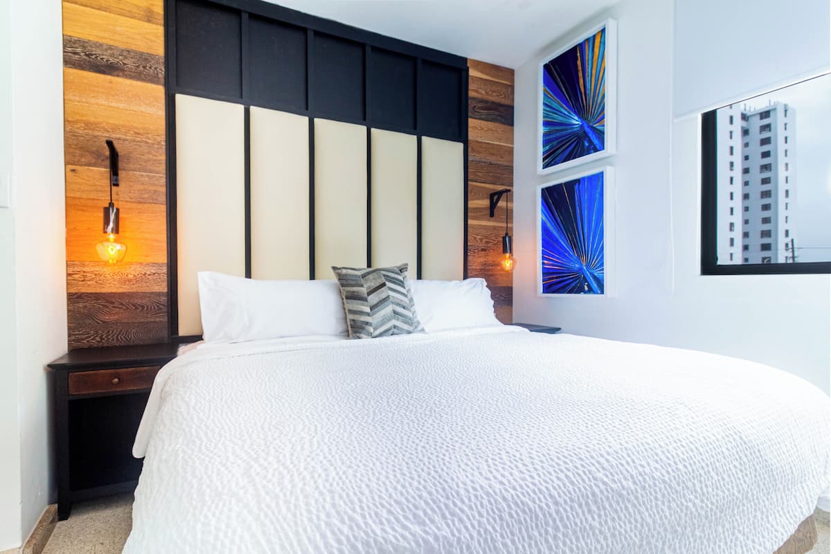 A hotel room with a large bed, wooden panel wall, two wall-mounted lights, and a window with a view of a building.