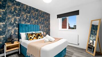 Un dormitorio moderno con una cama grande, un cabecero azul, una mesita de noche de madera, una mesilla auxiliar, un espejo y una ventana con vistas a edificios.