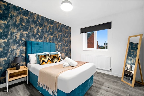 A modern bedroom with a large bed, a blue headboard, a wooden nightstand, a bedside table, a mirror, and a window with a view of buildings.