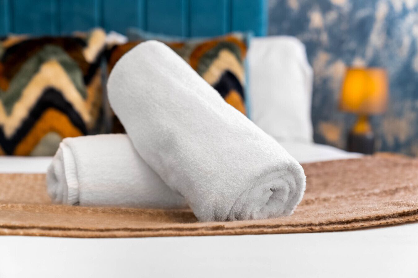 Una toalla blanca doblada con cuidado sobre un felpudo marrón, un cabecero tapizado en azul y una almohada estampada al fondo.