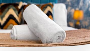 Una toalla blanca doblada con cuidado sobre un felpudo marrón, un cabecero tapizado en azul y una almohada estampada al fondo.