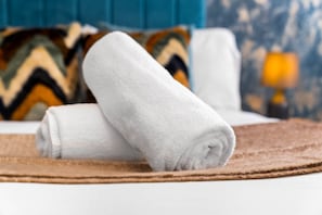 A neatly folded white towel on a brown mat, a blue upholstered headboard, and a patterned pillow in the background.