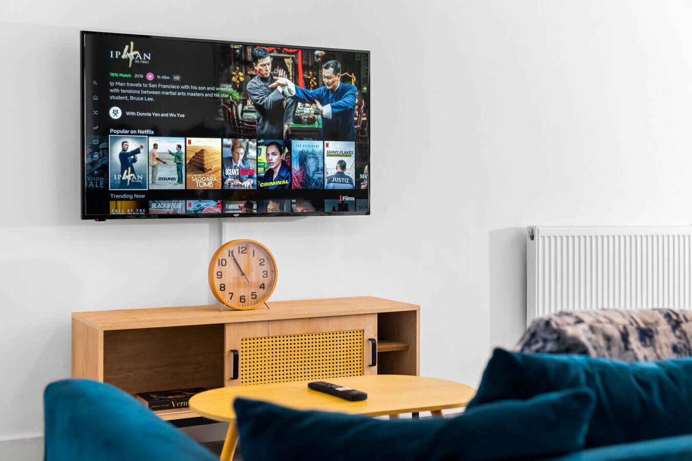 Una sala de estar moderna con un televisor de pantalla plana montado en la pared mostrando una película. Debajo del televisor, hay una mesa de centro de madera con un reloj y un control remoto.