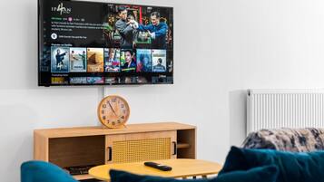 Una sala de estar moderna con un televisor de pantalla plana montado en la pared mostrando una película. Debajo del televisor, hay una mesa de centro de madera con un reloj y un control remoto.