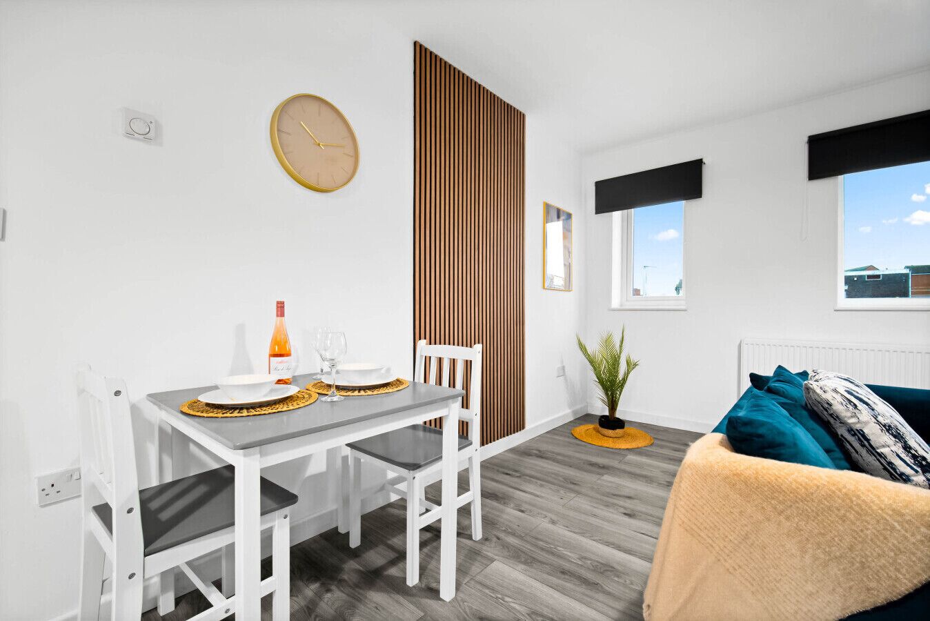 Una habitación moderna y bien iluminada con una mesa de comedor dispuesta para dos personas, una silla blanca, un sofá con cojines color turquesa, una separador de listones de madera, una planta en maceta y un reloj de pared.
