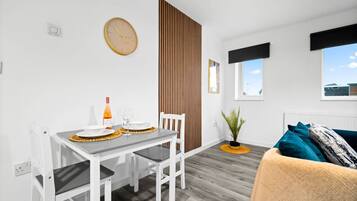 Una habitación moderna y bien iluminada con una mesa de comedor dispuesta para dos personas, una silla blanca, un sofá con cojines color turquesa, una separador de listones de madera, una planta en maceta y un reloj de pared.