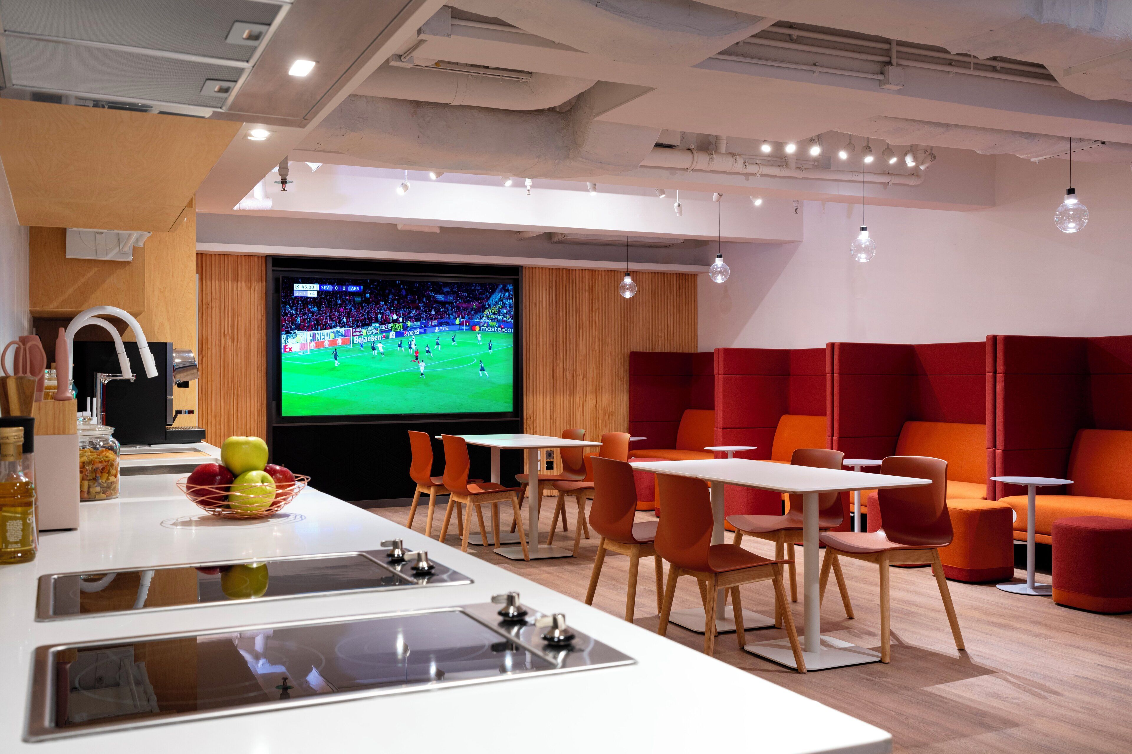 A modern lounge area with a large TV screen displaying a sports event, seating arrangements, and a kitchen counter with a fruit bowl.