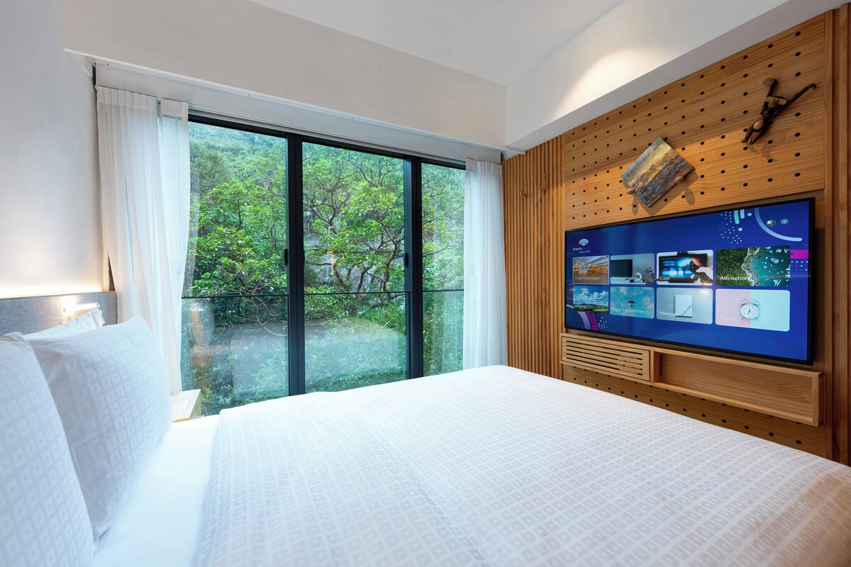 A modern hotel room with a large bed, a TV mounted on a wooden wall, and a sliding glass door leading to a balcony with a view of greenery.