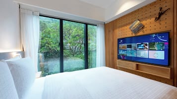 Ein modernes Hotelzimmer mit einem großen Bett, einem an einer Holzwand montierten Fernseher und einer Glastür, die auf einen Balkon mit Blick auf Grünflächen führt.