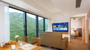 Habitación de hotel moderna con zona de comedor, sofá, televisor y vistas a un paisaje montañoso.