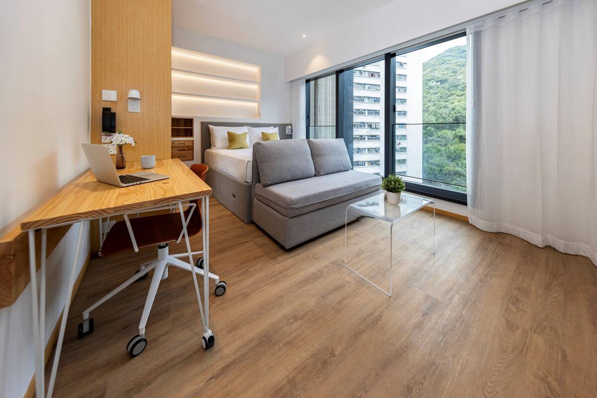 A modern hotel room with a wooden desk, a grey sofa, a small white table, and a large window offering a view of a building and greenery.