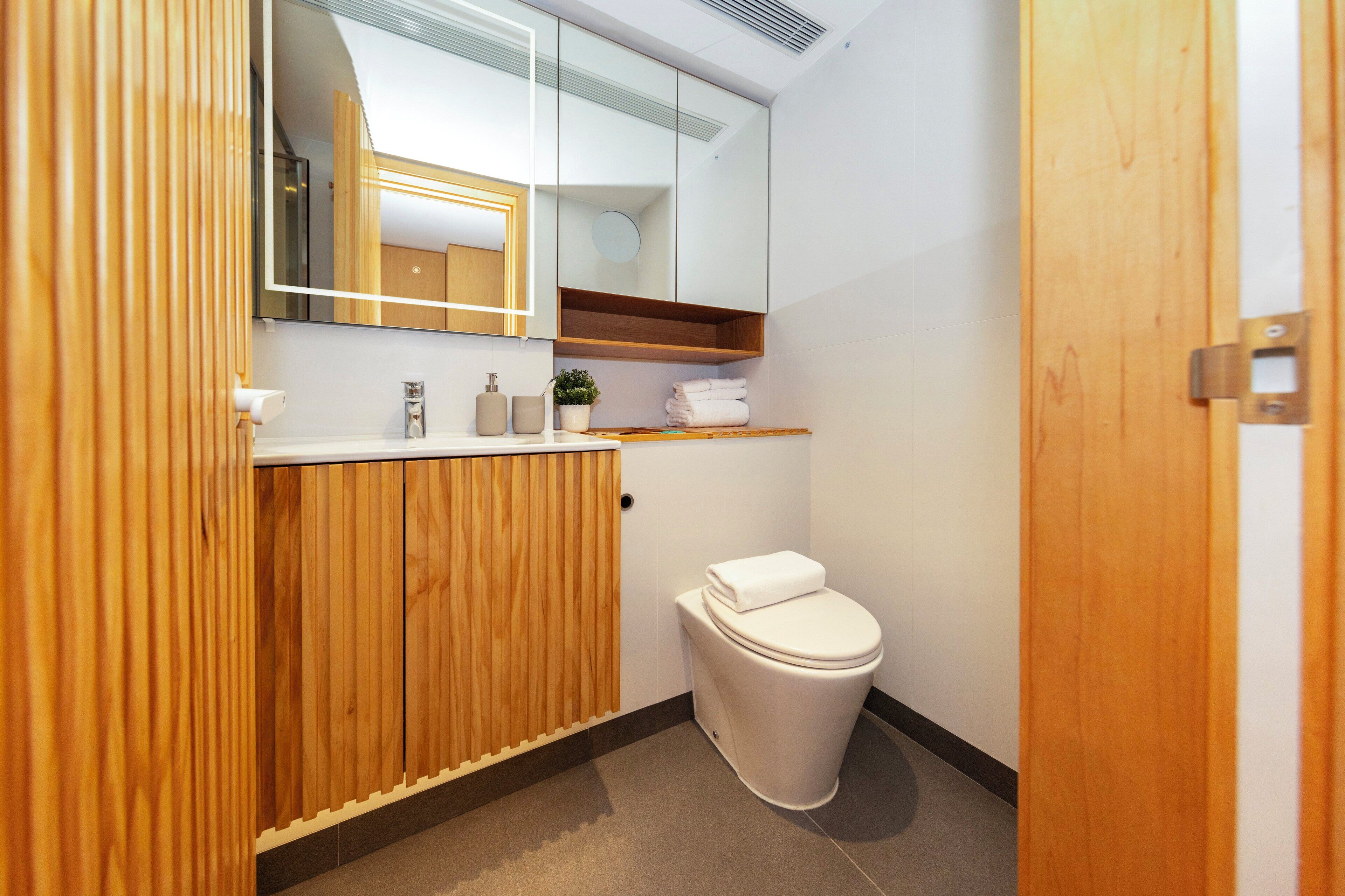 A modern bathroom with a wooden vanity, a toilet, a mirror, and a small shelf with towels.