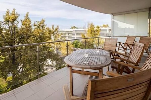 A balcony with a round table and wooden chairs, overlooking a cityscape with trees and buildings.