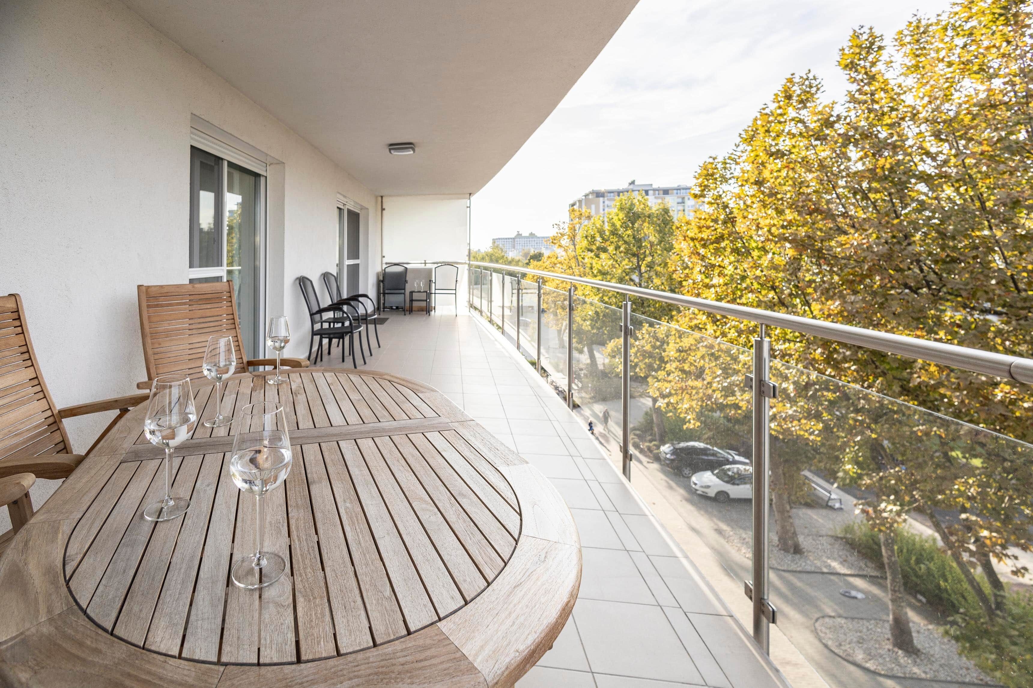 A round wooden table with chairs and wine glasses on a balcony with a glass railing.