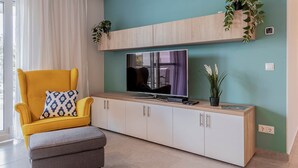 A living room with a yellow armchair, a grey ottoman, a television on a wooden console, and potted plants. - LB - Garden Pearl II Siófok (Siófok)