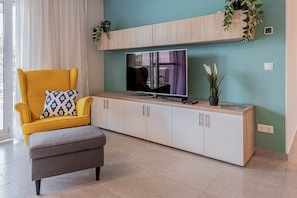 A living room with a yellow armchair, a grey ottoman, a television on a wooden console, and potted plants. - LB - Garden Pearl II Siófok (Siófok)