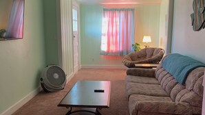 A living room with a brown carpet, a brown sofa, a small table, a fan, and a window with curtains.