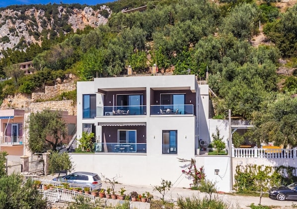 Un edificio moderno de dos pisos con balcones, rodeado de vegetación y una ladera rocosa.