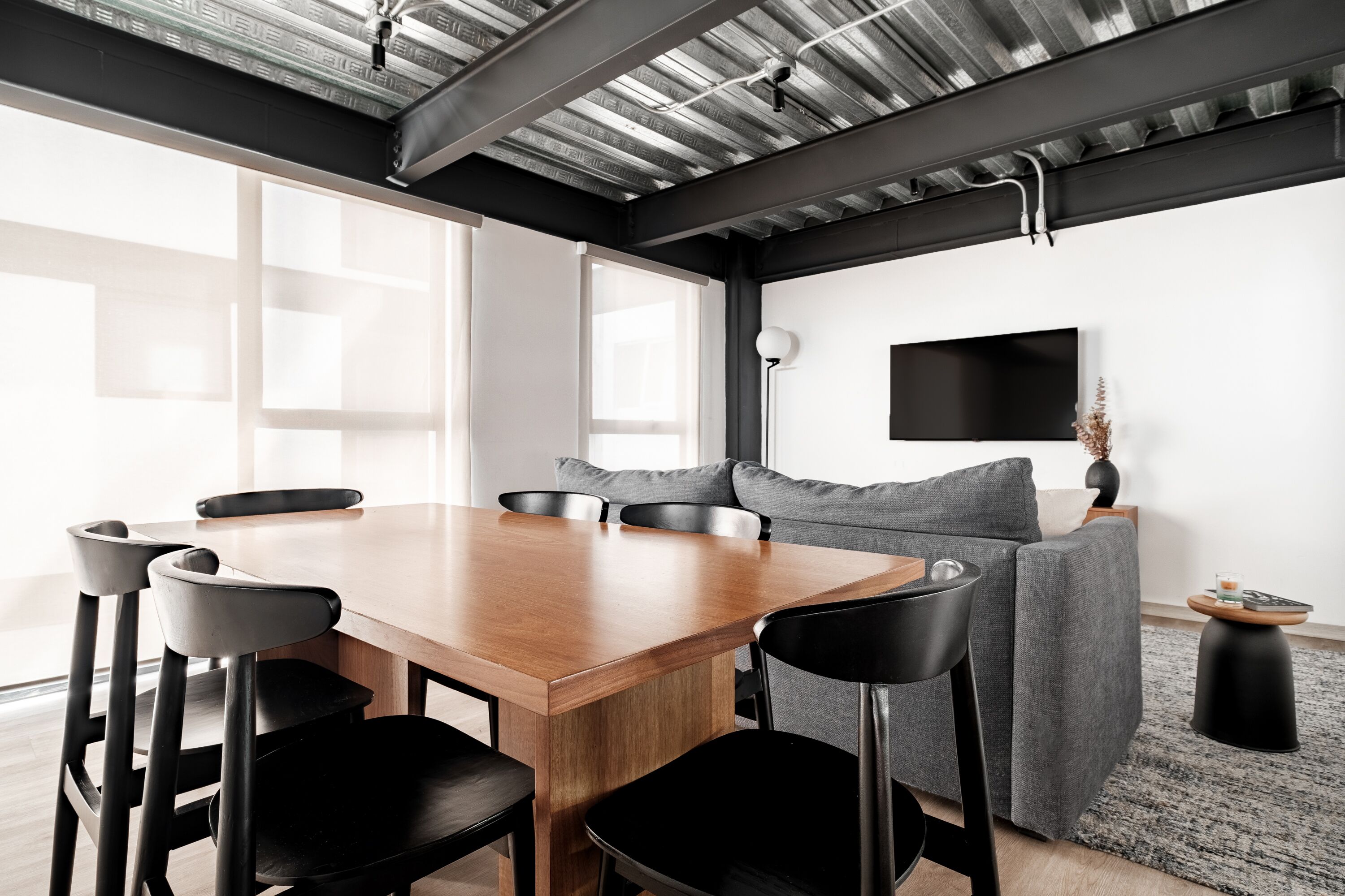 A modern living room with a wooden dining table, black chairs, a grey sofa, and a flat-screen TV mounted on the wall.