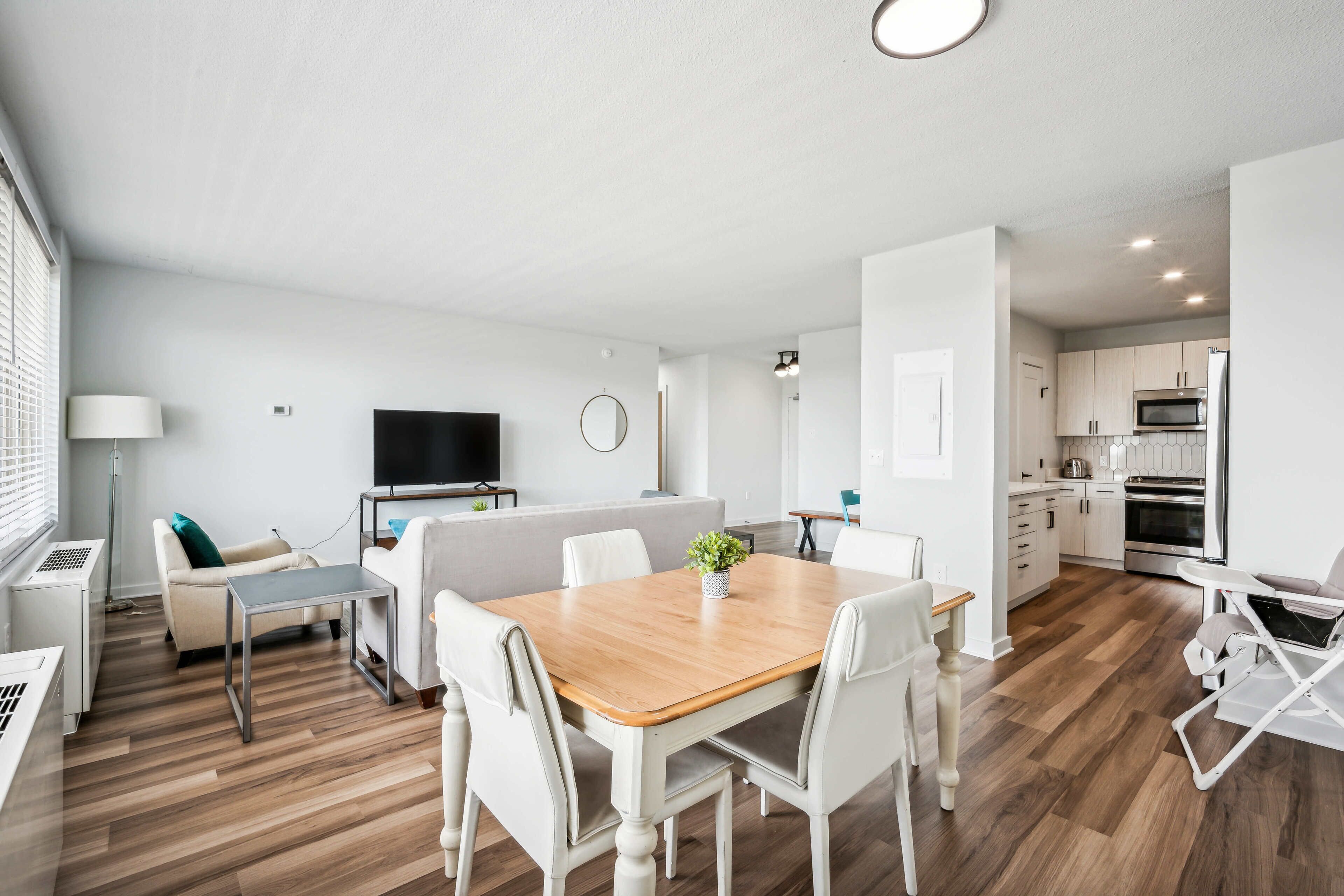 A modern, well-lit living room with a wooden dining table, white chairs, a sofa, and a TV.