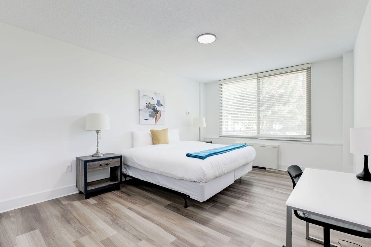 Een moderne slaapkamer met een bed, nachtkastjes, een bureau en een raam met jaloezieën.