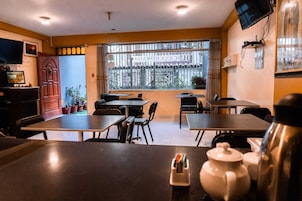 Um restaurante com mesas e cadeiras, uma televisão na parede e uma janela com vista para edifícios.