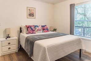 Un dormitorio con cama, mesita de noche, lámpara, un cuadro en la pared y ventana con vistas a árboles.