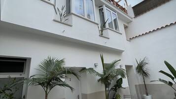Un bâtiment à deux étages, doté de nombreuses fenêtres et d’un balcon. On peut voir plusieurs plantes en pot dans la cour.