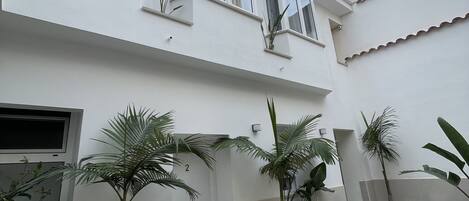 Un edificio de dos pisos con múltiples ventanas y un balcón. Varias plantas en macetas decoran el patio.