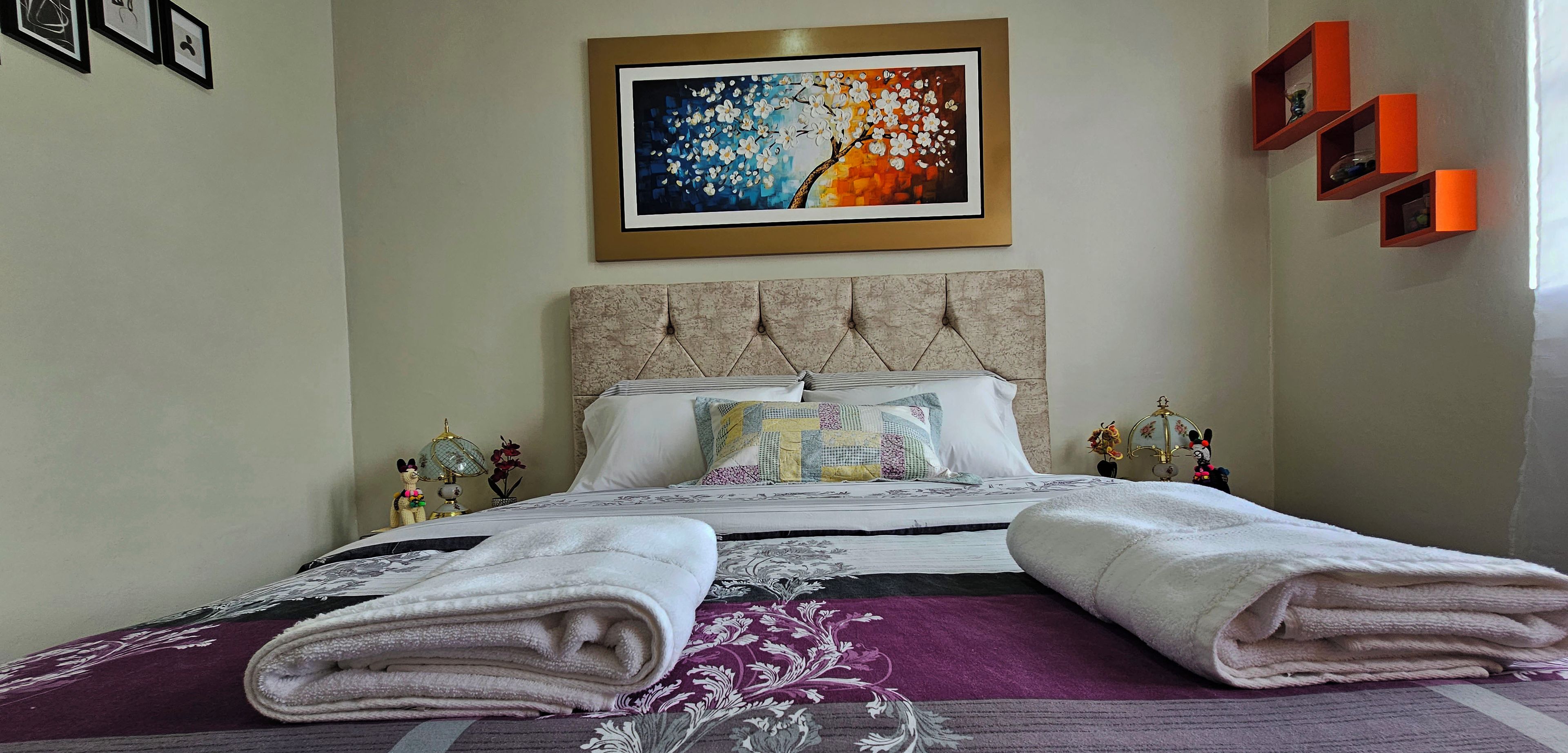 Een net opgemaakt bed met opgevouwen handdoeken, een decoratief kussen en een kleurrijk schilderij erboven.