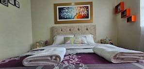 A neatly made bed with folded towels, a decorative pillow, and a colorful painting above it.