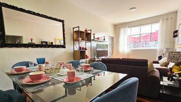 Een eetkamer met een glazen tafel voor vier personen, omringd door blauwe stoelen. Achtergrond: een bank en een televisie.