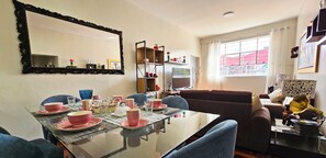 A dining room with a glass table set for four, surrounded by blue chairs. There is a sofa and a television in the background.
