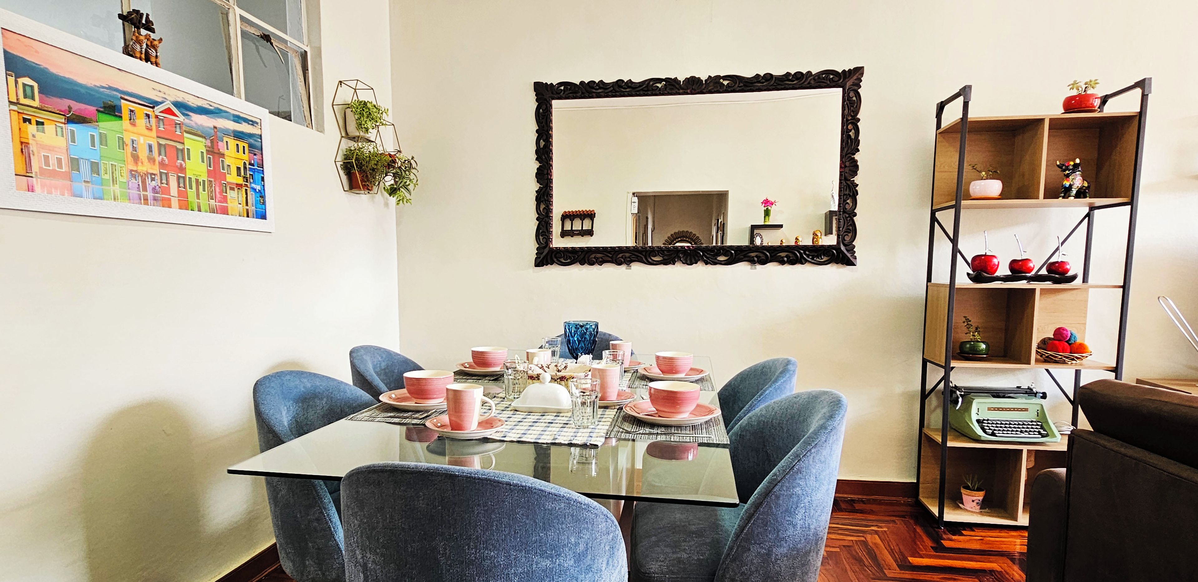 Een eetkamer met een glazen tafel, blauwe stoelen en roze servies.