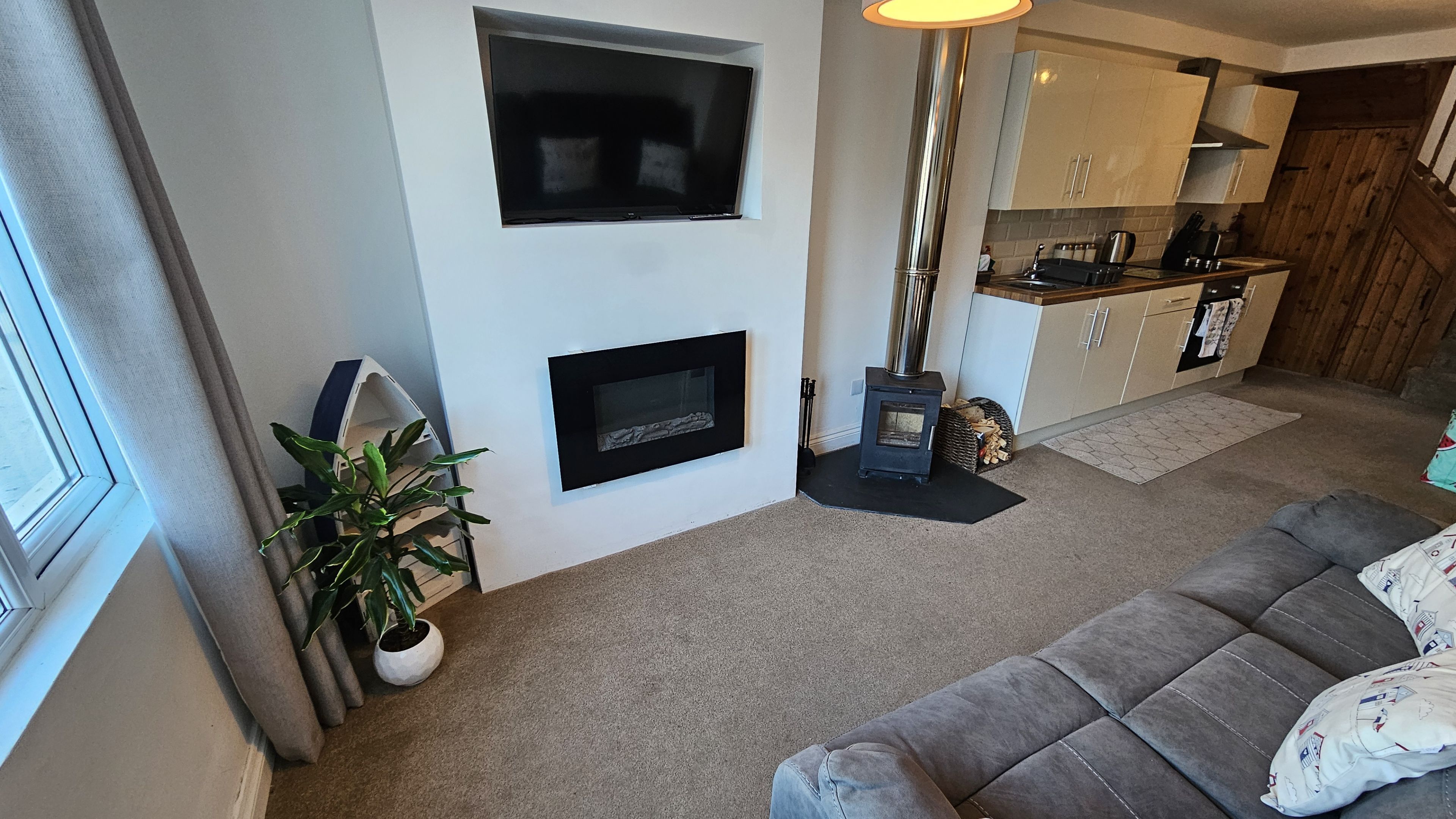 A living room with a sofa, a fireplace, a TV mounted on the wall, and a kitchen area with cabinets and a stove.