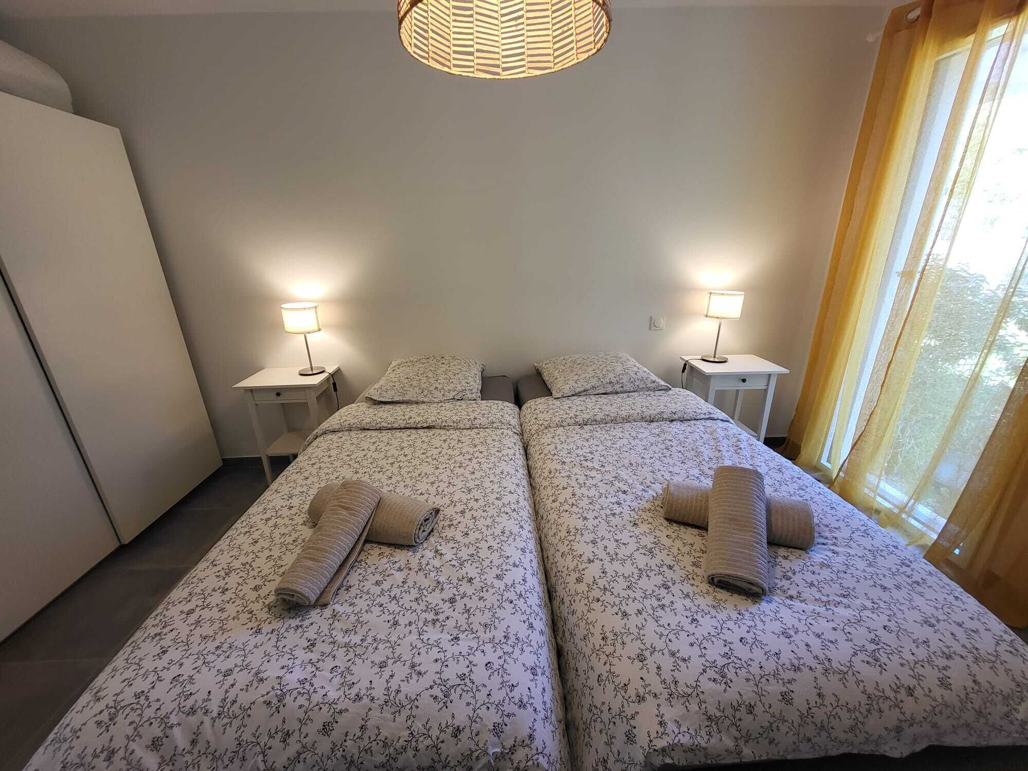 Hotelkamer met twee bedden, elk met een bloemenprintdeken en een opgerolde handdoek. Er staan nachtlampjes, er is een raam met gordijnen en een hanglamp.