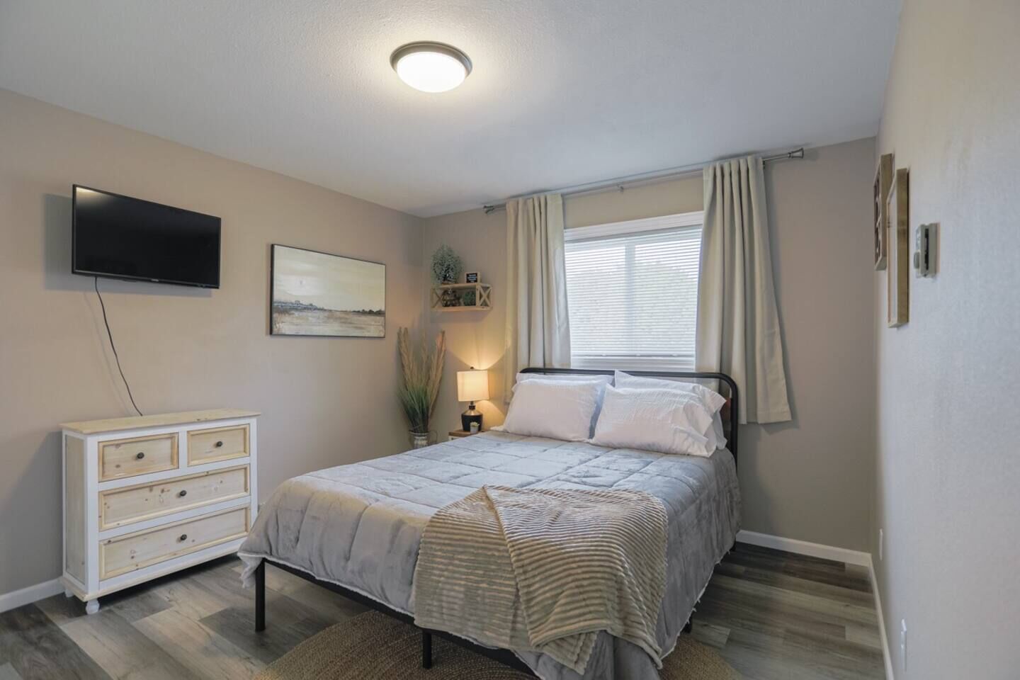 Une chambre à coucher comprenant un lit, une télévision, une commode, une fenêtre avec des rideaux et un luminaire au plafond.