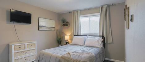 Une chambre à coucher comprenant un lit, une télévision, une commode, une fenêtre avec des rideaux et un luminaire au plafond.