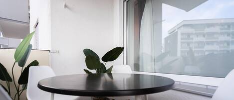 A small balcony with a round table and two white chairs, a potted plant, and a view of a building through a window.