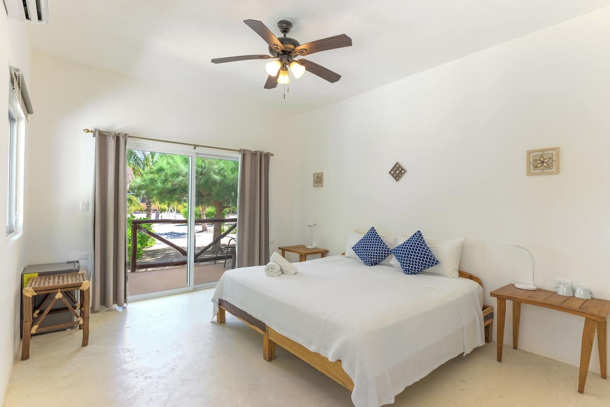 Um quarto com cama, ventilador de teto, uma mesa pequena e janela com cortinas.
