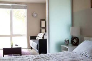 Ein Schlafzimmer mit einem Bett, einem Nachttisch, einer Lampe, einer Uhr, einem Sofa und einem Fenster mit Jalousien.