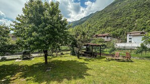 Un espacio al aire libre con césped, un banco de madera, una mesa de picnic y un pabellón. Se ven casas al fondo con un paisaje montañoso.