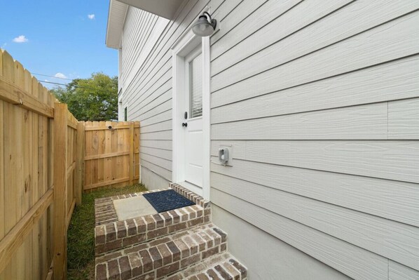 A house with a wooden fence, a brick staircase leading to a white door, and a blue mat at the door's entrance. - Bright & open/walk to downtown! (Pensacola)
