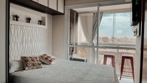 Une chambre avec un grand lit, une vue sur la ville depuis un balcon et un tabouret rouge à l’extérieur.