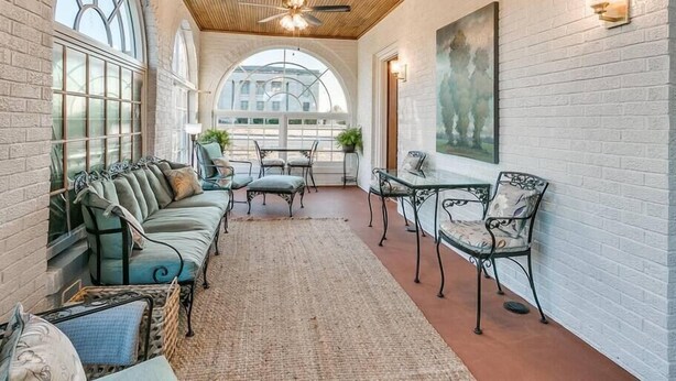 A sunlit room with a wooden ceiling, large windows, a sofa, chairs, and a painting on the wall.