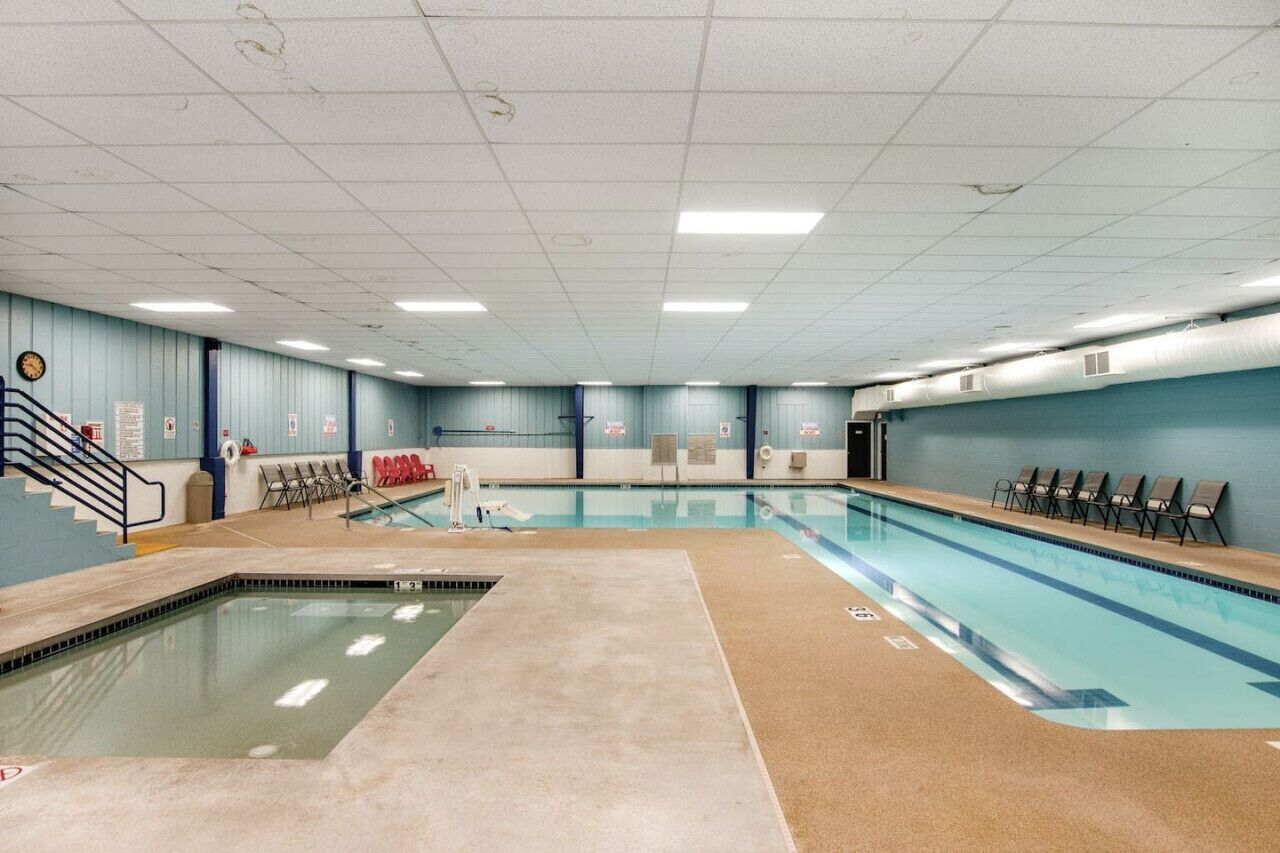 Ein großes Hallenbad mit Fliesenboden, einer Treppe links und einer Reihe von Stühlen an der rechten Wand.