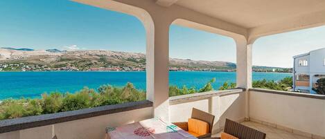 A balcony with a table and chairs, overlooking a coastal view with a clear blue sea and distant hills.