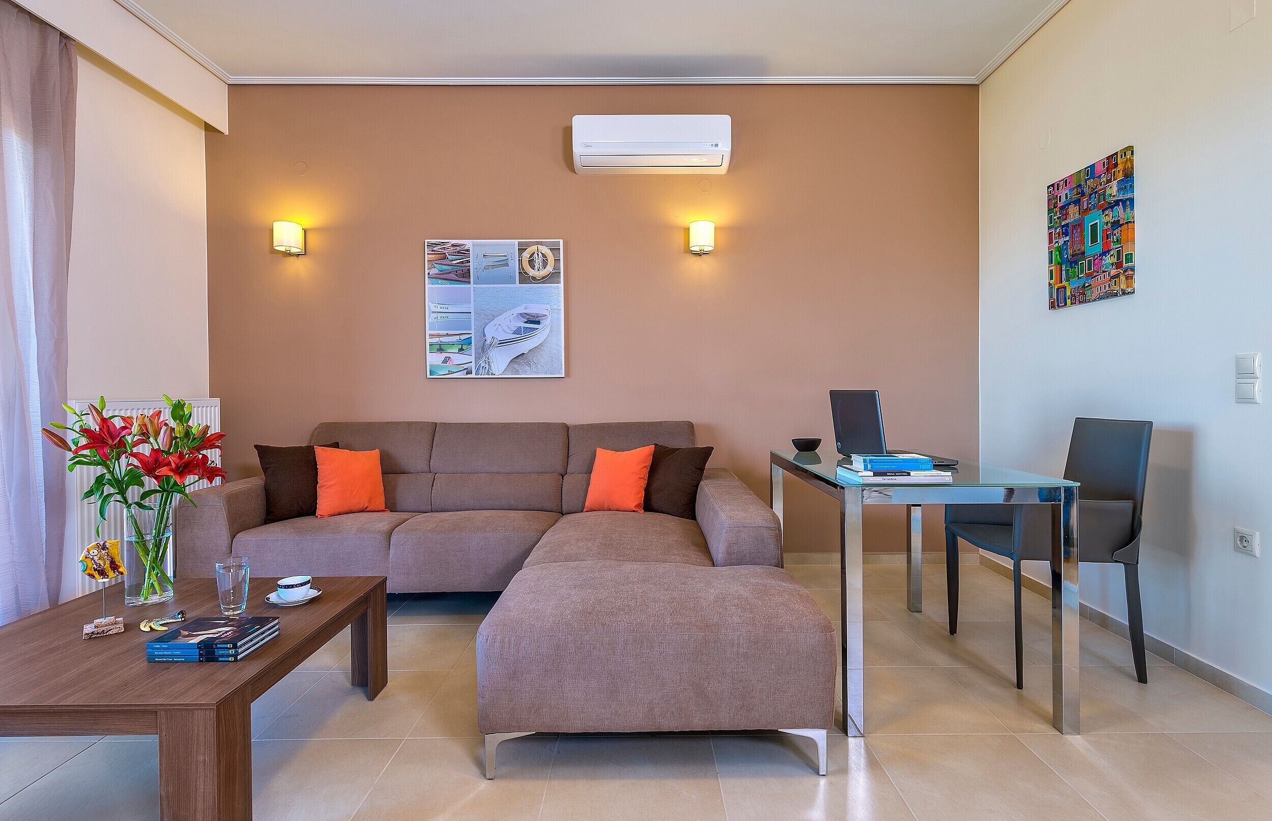 A modern living room with a sectional sofa, a glass coffee table, a side table with books and a vase of flowers, a small table with a laptop, and a colorful artwork on the wall.