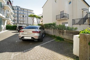 A residential street with a parked car, a building with balconies, and a concrete planter with greenery.